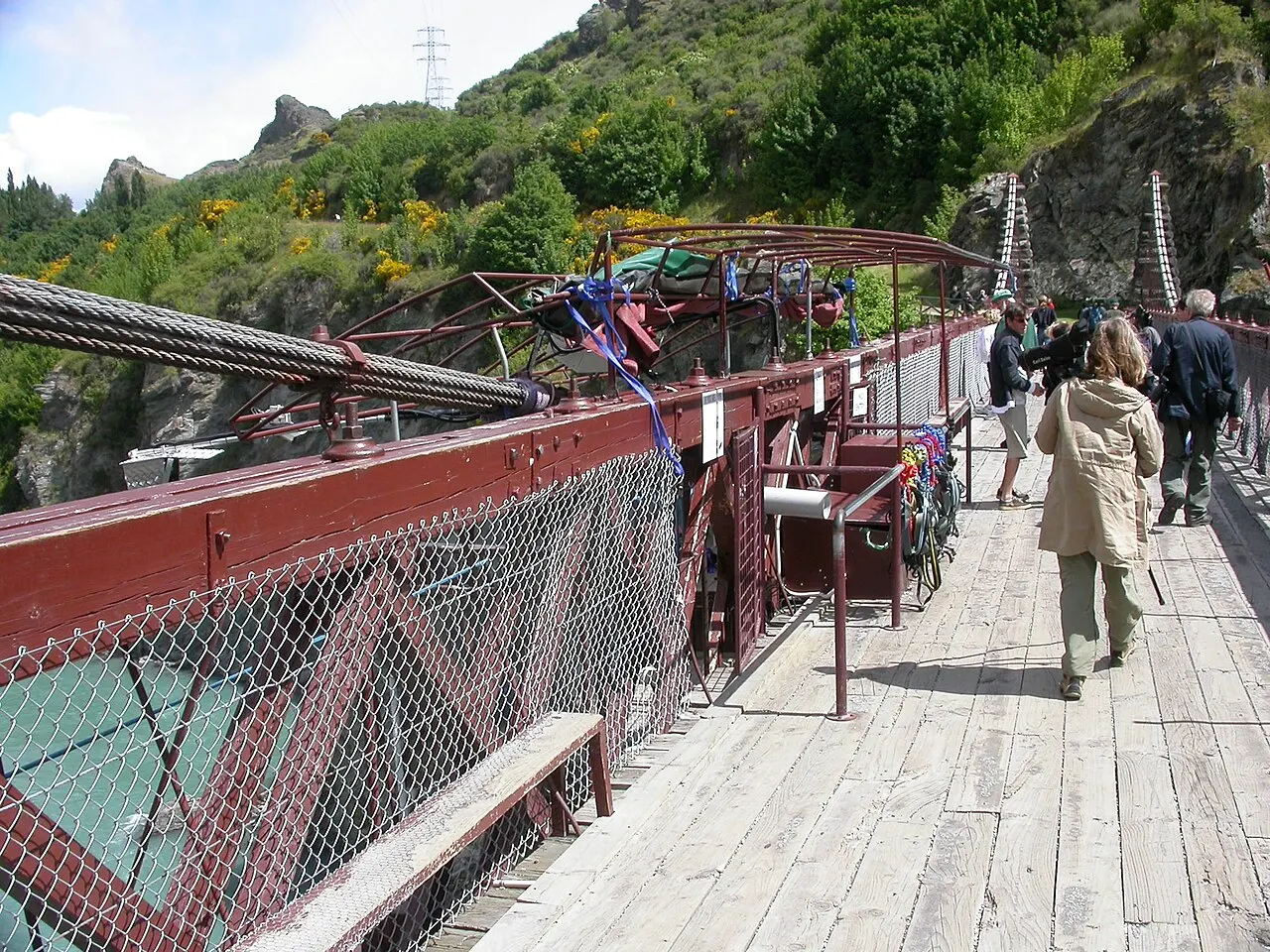 Plataforma de salto na Ponte do Kawarau, com estrutura de metal vermelha, redes de proteção nas laterais e pessoas caminhando pelo deque de madeira.