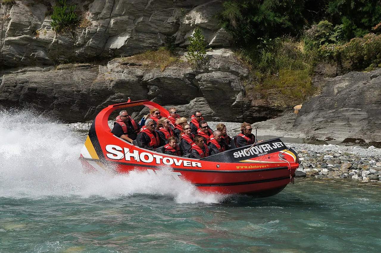 Lancha vermelha do Shotover Jet em alta velocidade rente às rochas do cânion, com passageiros a bordo.