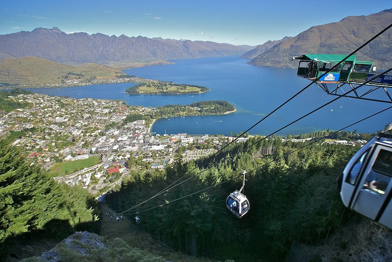 Cabines do teleférico Skyline Gondola subindo a montanha, com vista aérea de Queenstown, o lago Wakatipu e cordilheiras ao fundo, sob céu claro.