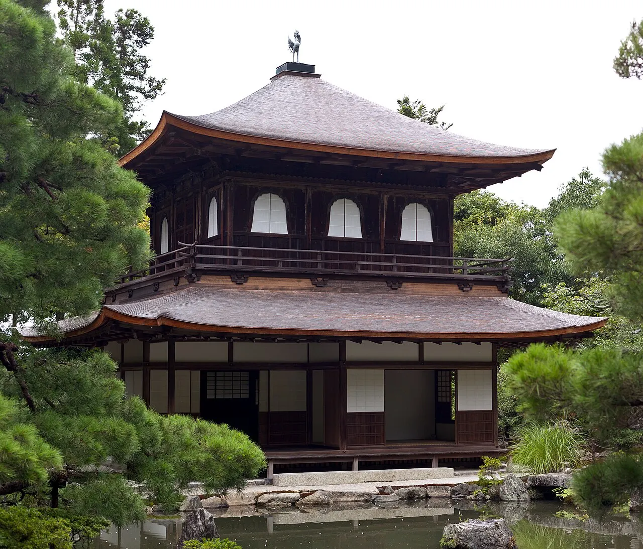 Pavilhão de dois andares em madeira escura e telhado claro, entre pinheiros podados.