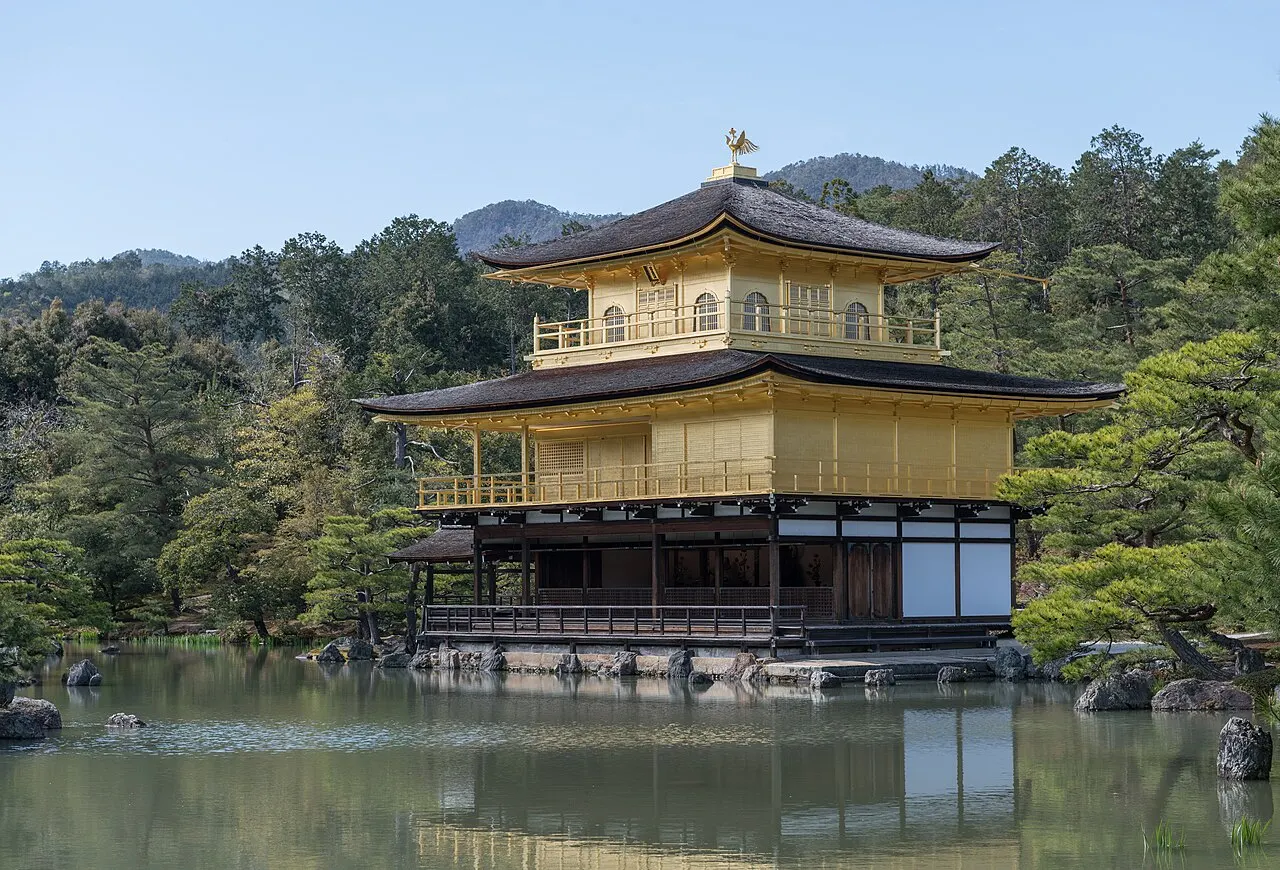 Pavilhão de três andares coberto de folha de ouro, à beira de um lago, com pinheiros atrás.