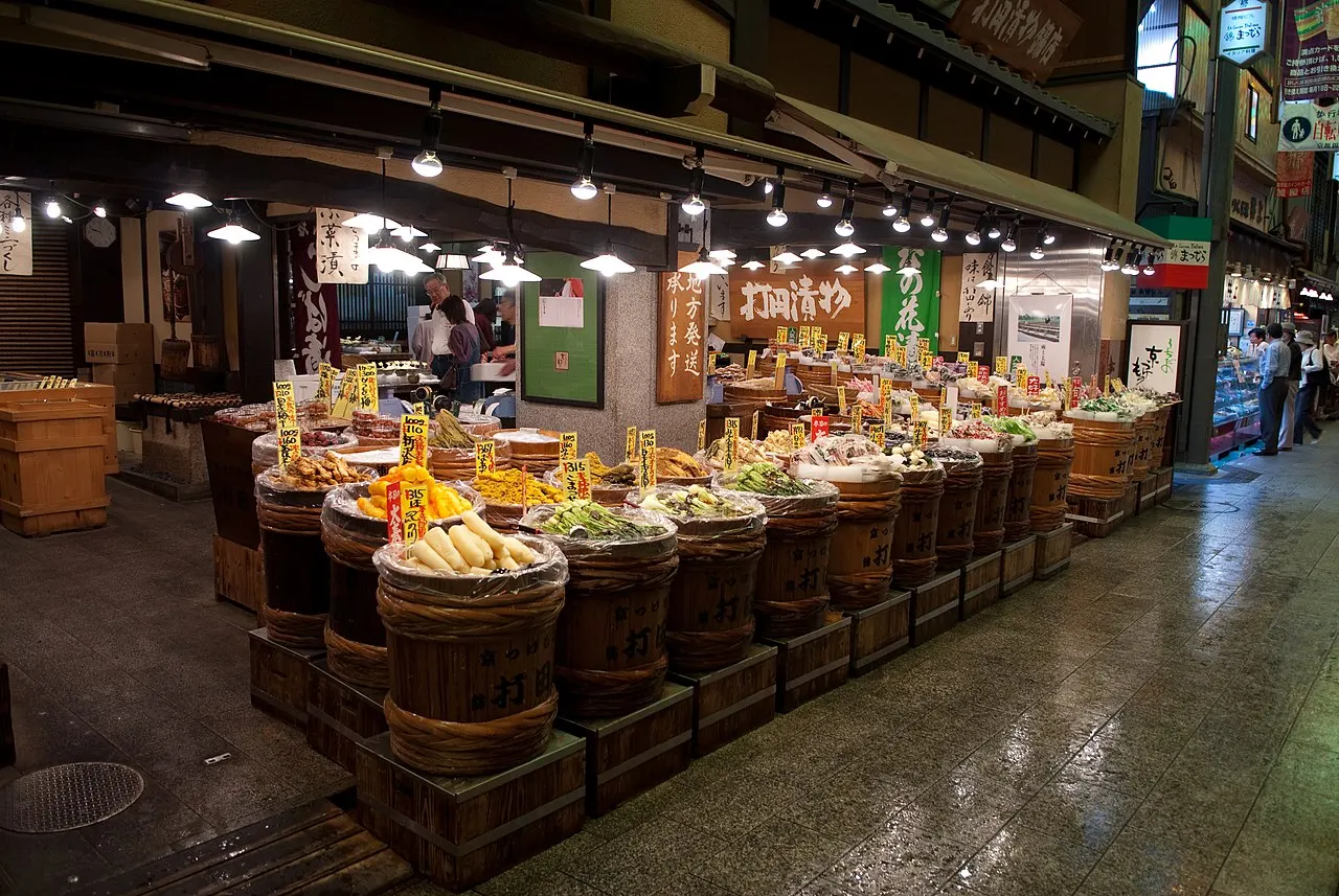 Fileiras de barris de madeira com conservas coloridas numa banca do mercado coberto.