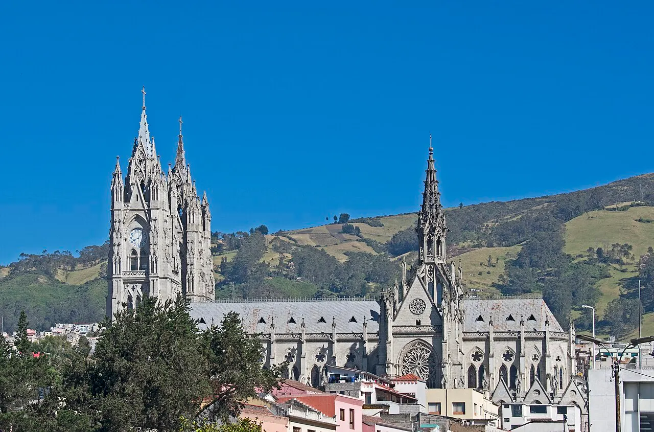 Torres neogóticas em pedra clara da Basílica del Voto Nacional, com pináculos esguios e rosácea, encostadas numa colina verde sob céu azul.