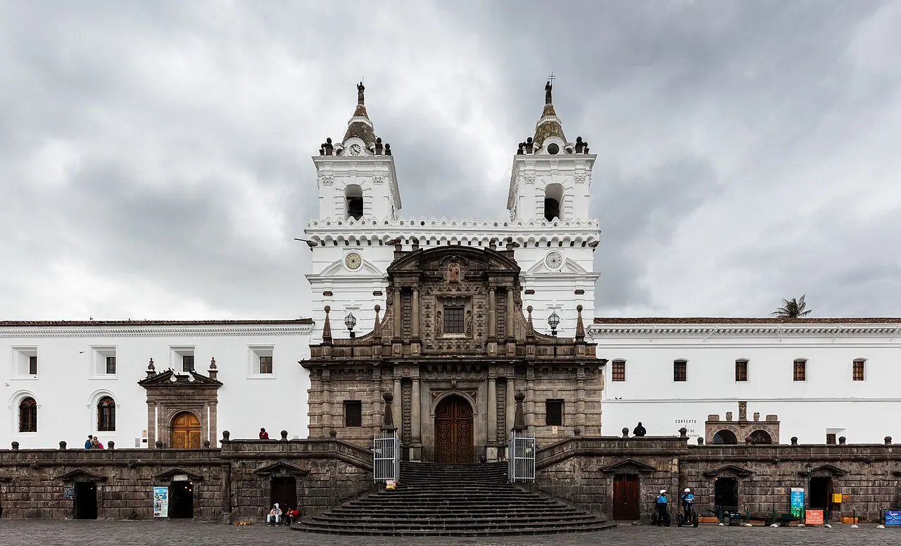 Fachada branca da igreja de San Francisco, com duas torres sineiras, portal de pedra esculpida e uma ampla escadaria descendo até a praça, sob céu nublado.