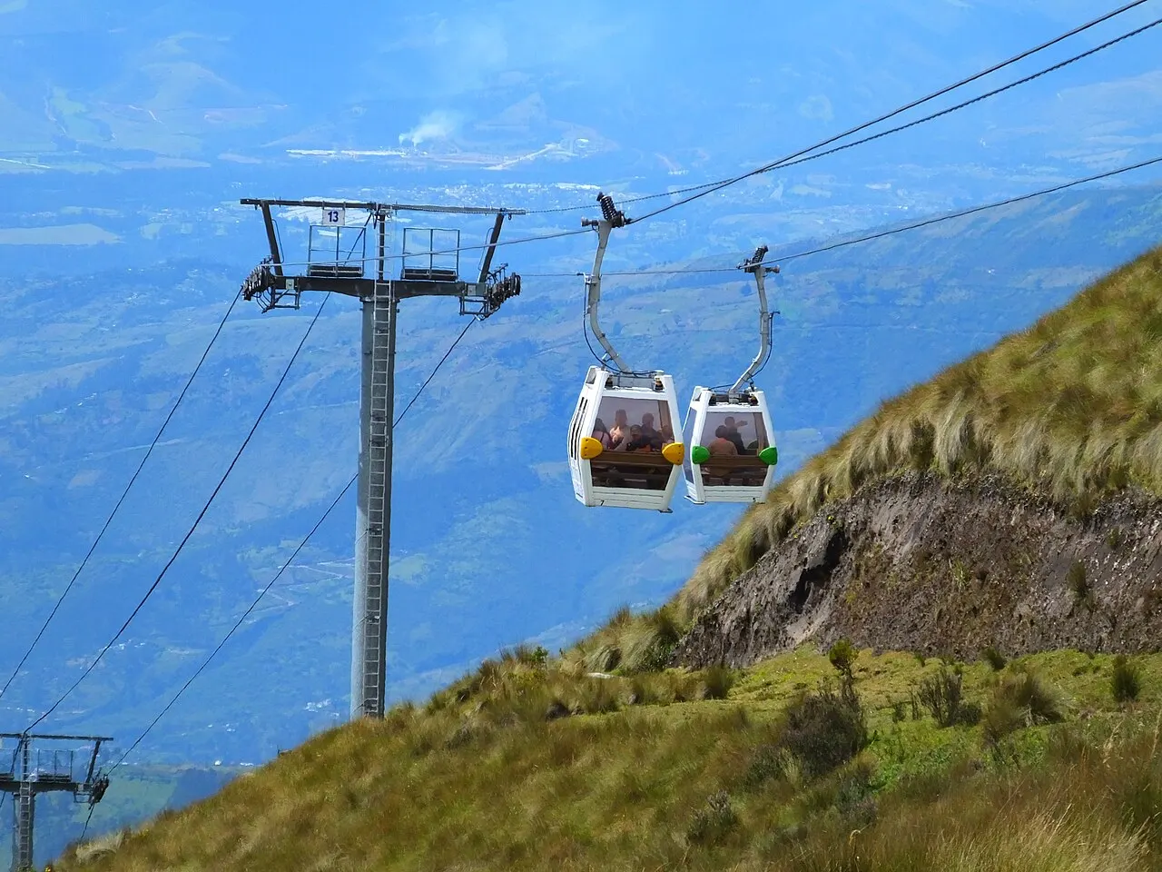 Duas cabines brancas do TelefériQo cruzando junto a uma torre metálica, sobre a encosta de capim amarelo do Pichincha, com o vale e as montanhas azuladas de Quito ao fundo.