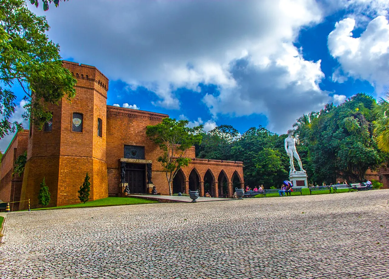 O prédio em forma de castelo do Instituto Ricardo Brennand, de tijolo aparente com torre e arcadas, diante de um pátio de paralelepípedos com uma estátua branca.
