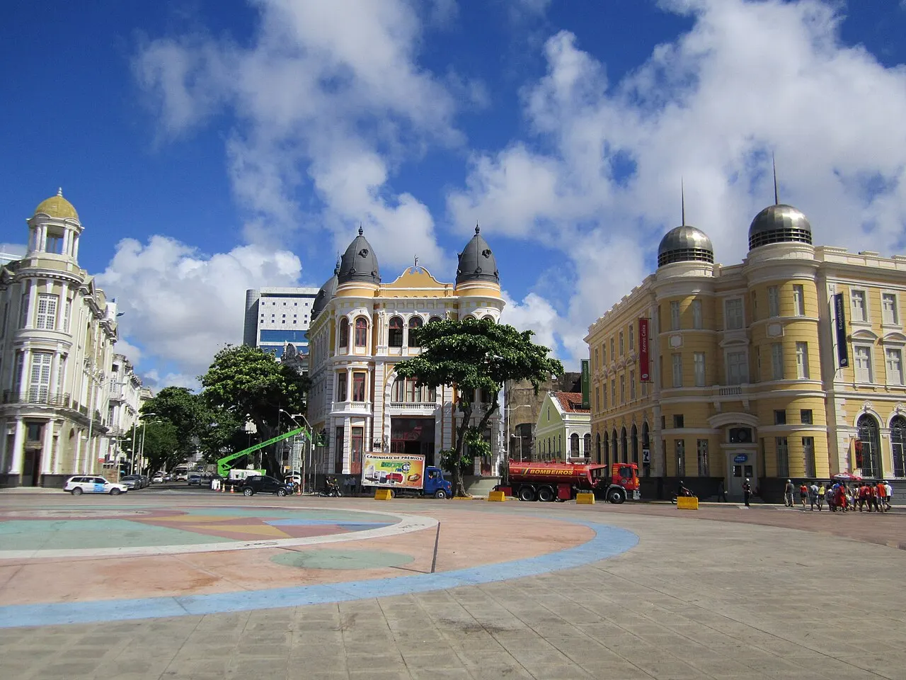 A Praça Rio Branco, no Recife Antigo, com o piso desenhado em curvas coloridas e prédios ecléticos de cúpula em volta, sob céu com nuvens.