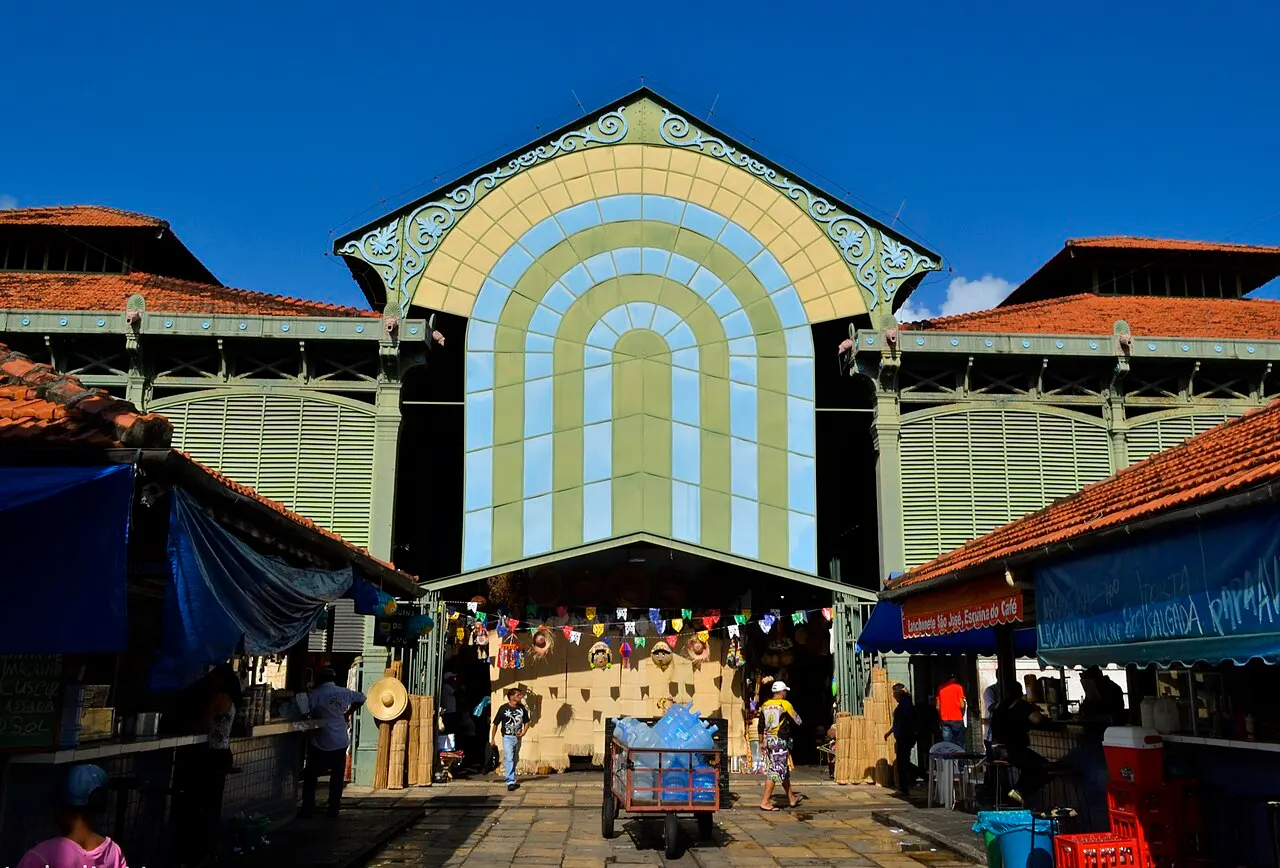 La façade du marché de São José, structure de fer verte avec une grande arche vitrée bleue et jaune, et des étals et des passants devant.