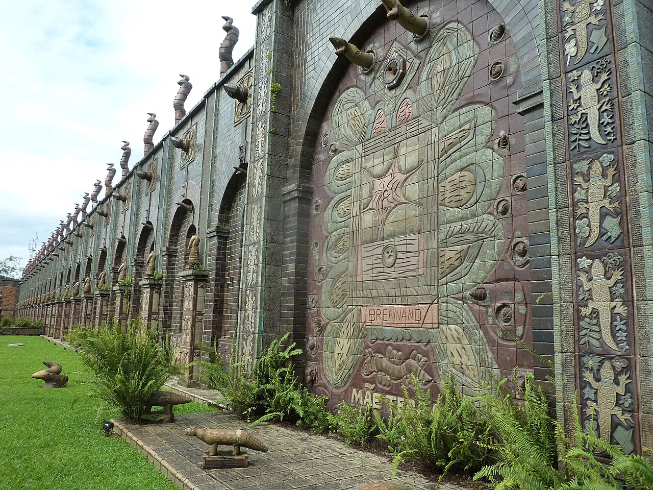 Um muro longo da Oficina Brennand coberto de relevos de cerâmica e encimado por pequenas esculturas, ao lado de um gramado.