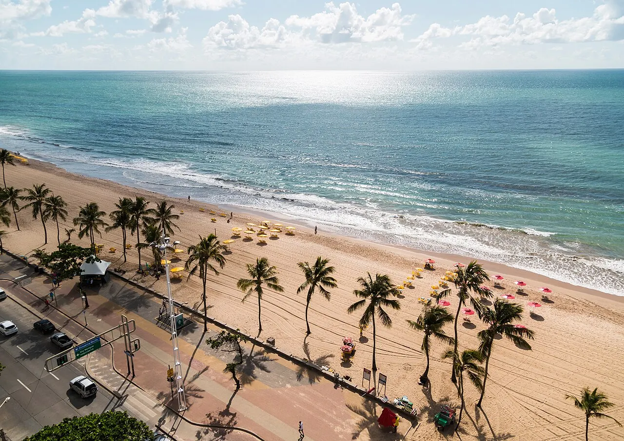 Vue en hauteur de la plage de Boa Viagem en 2018, avec des cocotiers sur la promenade, des parasols jaunes et roses sur le sable et la mer bleue au fond.