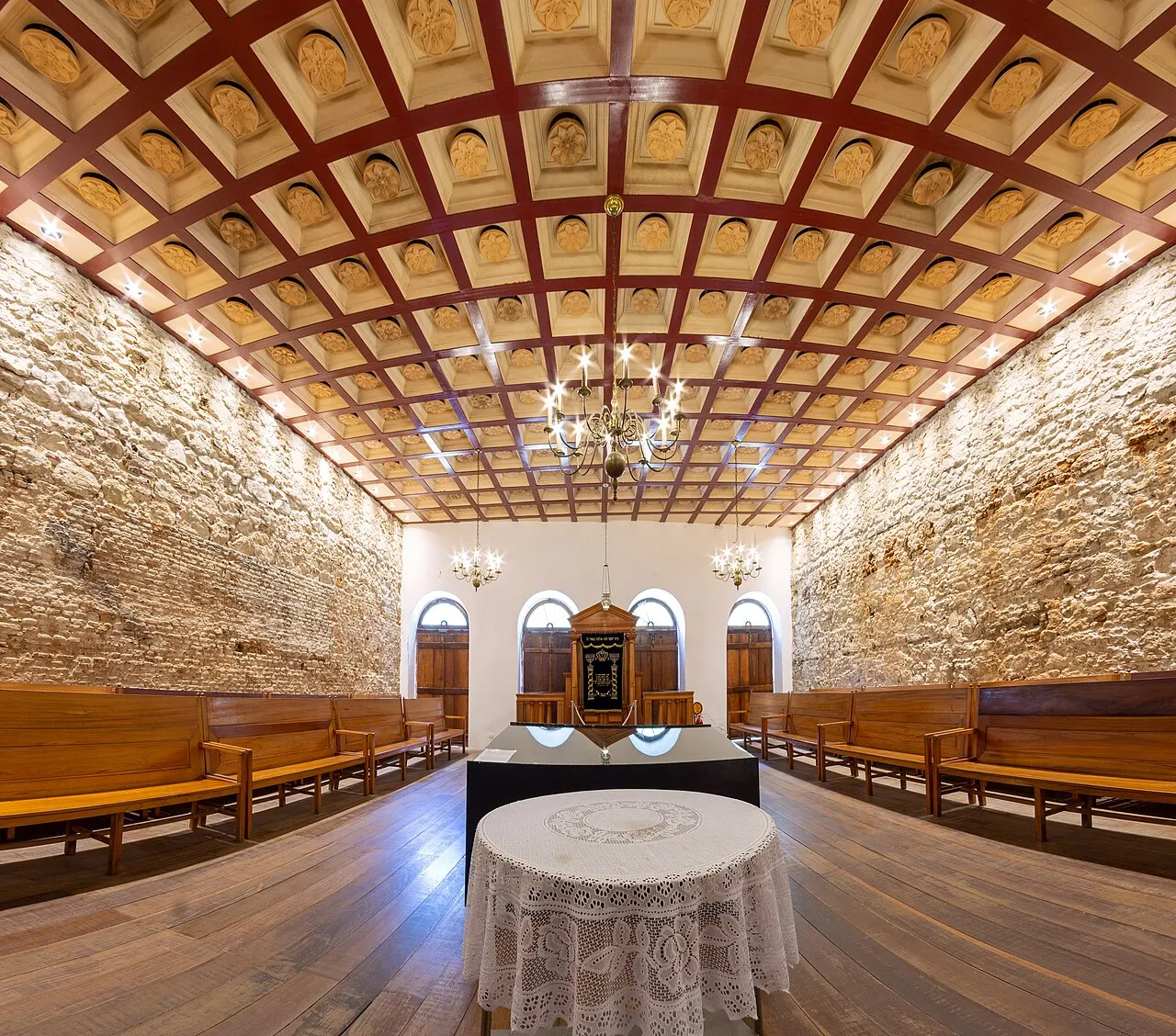 O salão da Sinagoga Kahal Zur Israel, com paredes de pedra aparente, teto de madeira em caixotões, bancos laterais e uma mesa ao centro.