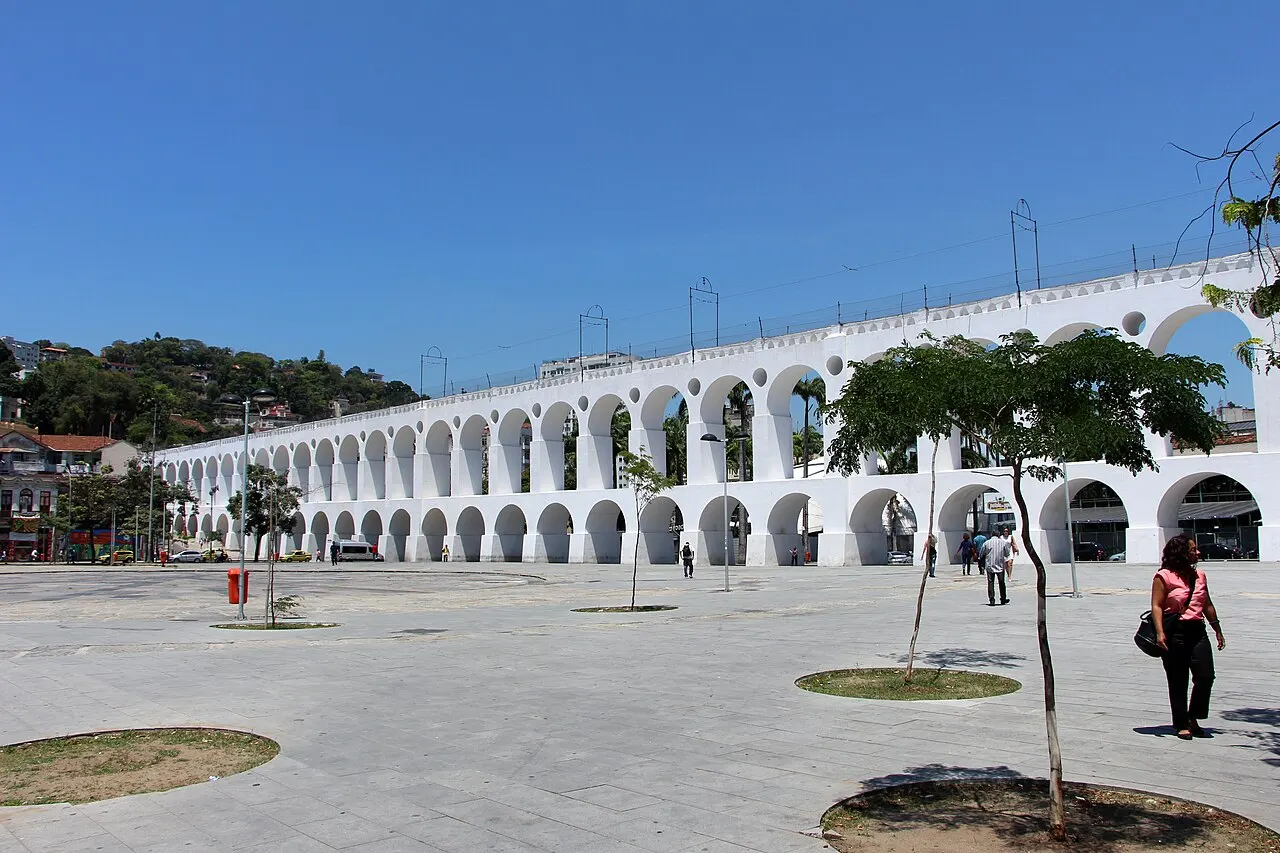 Os Arcos da Lapa, em fileira branca, vistos de uma praça ampla sob céu azul, com uma pessoa caminhando.
