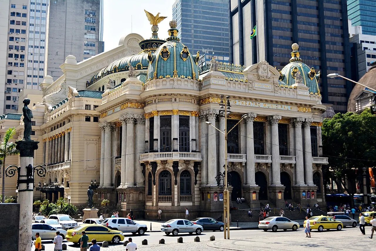 Fachada do Theatro Municipal do Rio de Janeiro, com cúpulas verdes e estátuas douradas, entre prédios altos.