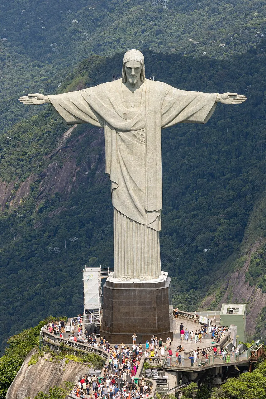 Estátua do Cristo Redentor de braços abertos sobre o pedestal, no alto do Corcovado, com a mata ao fundo.