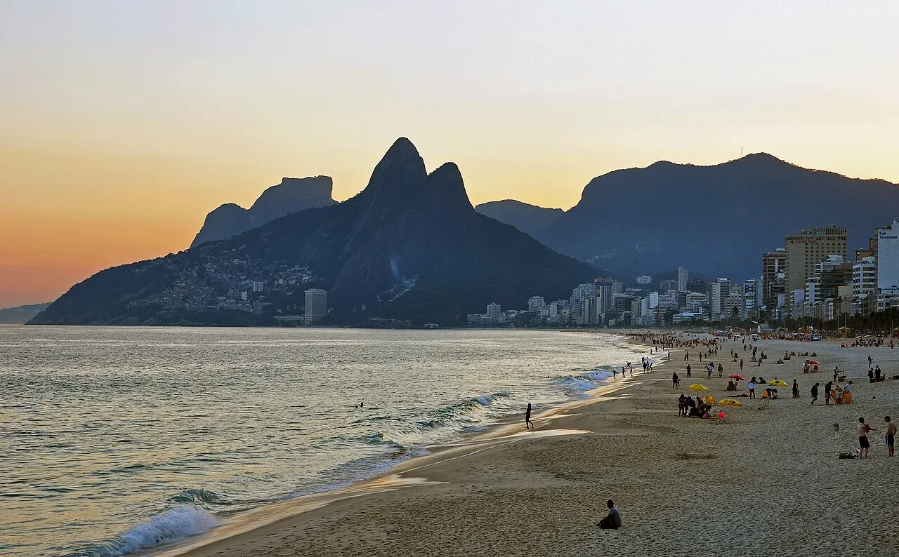 Praia de Ipanema ao entardecer, com as duas pontas do morro Dois Irmãos recortadas contra o céu alaranjado e gente na areia.