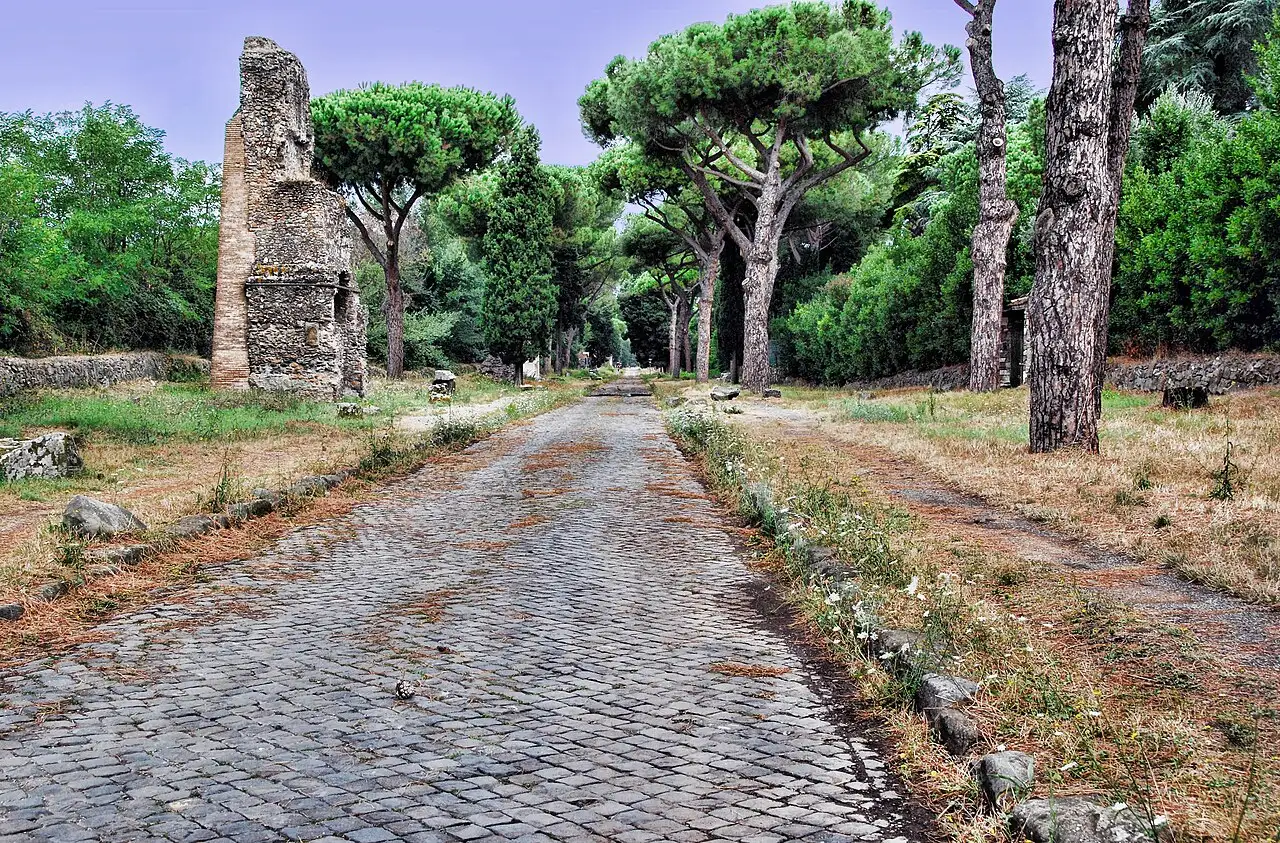 Un tronçon pavé de la Voie Appienne entre des pins, avec une ruine en brique au fond à gauche.