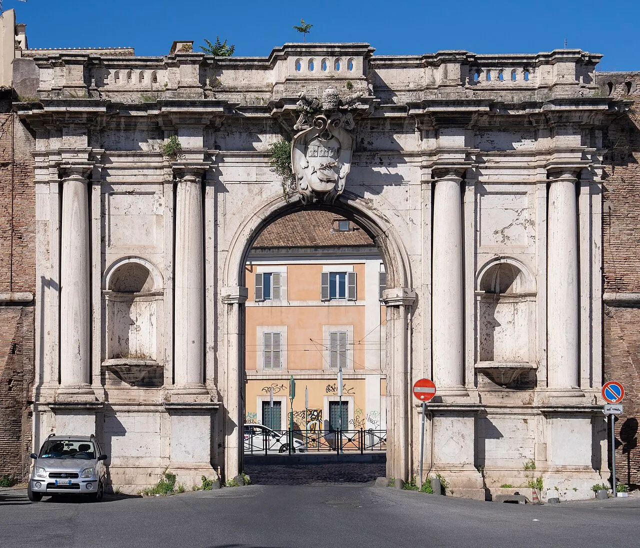 A Porta Portese, arco de pedra com colunas e frontão, vista de frente numa rua com carros estacionados.