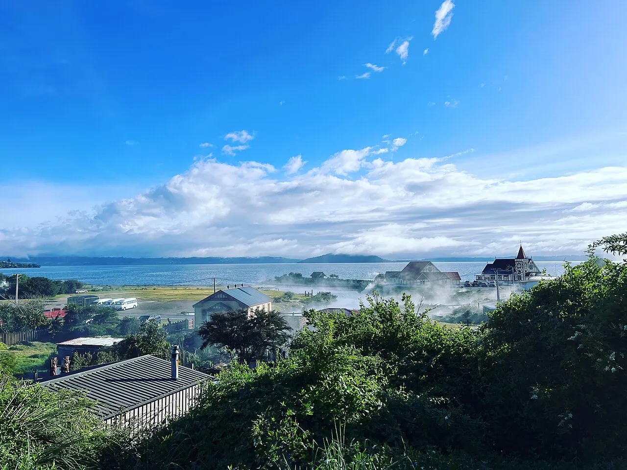 Vista do alto de casas à beira de um lago, com vapor subindo entre os telhados e a torre de uma igreja, e mata em primeiro plano.