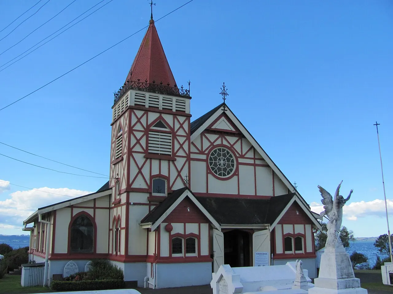 Igreja de madeira com enxaimel vermelho e creme, torre de telhado pontudo e janela redonda, com uma estátua de anjo branca ao lado.