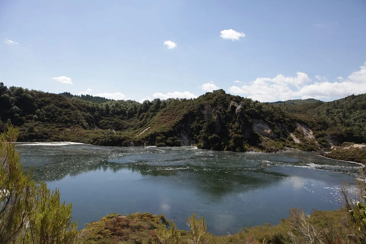 A crater lake with light steam on its surface, ringed by bush-covered slopes and a rock cliff, under a blue sky with clouds.