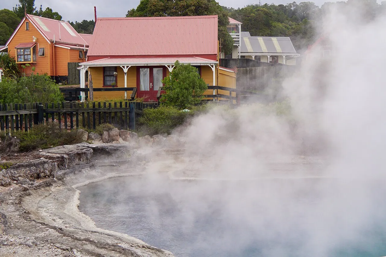 Wooden houses painted orange and yellow, with red roofs, behind a fence next to a hot pool giving off thick steam.
