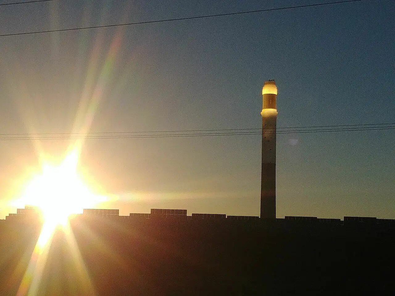 Torre solar alta contra o céu ao entardecer, com o receptor iluminado no topo e reflexo de luz à esquerda.