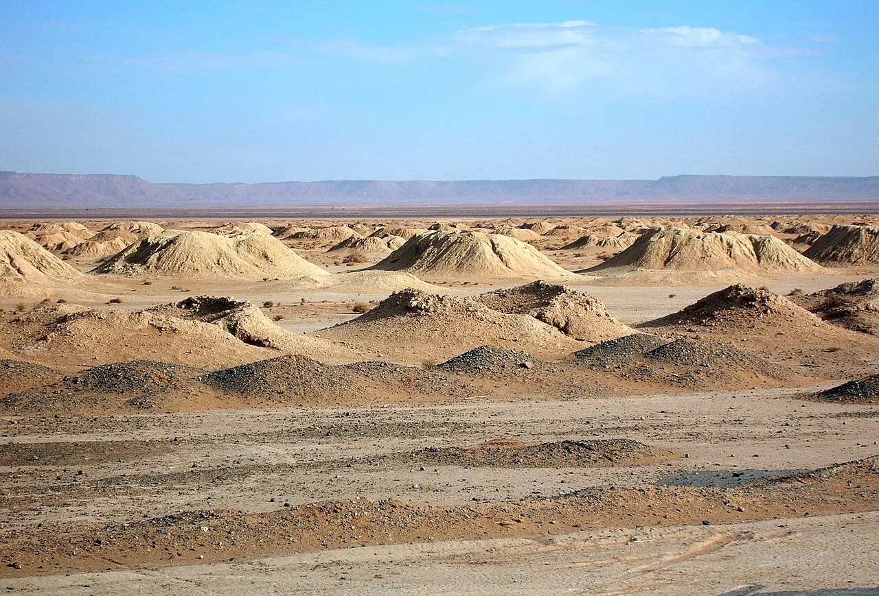 Fileiras de montes de terra clara alinhados sobre uma planície desértica plana, marcando os poços de acesso a um sistema de canais subterrâneos, com falésias ao fundo.