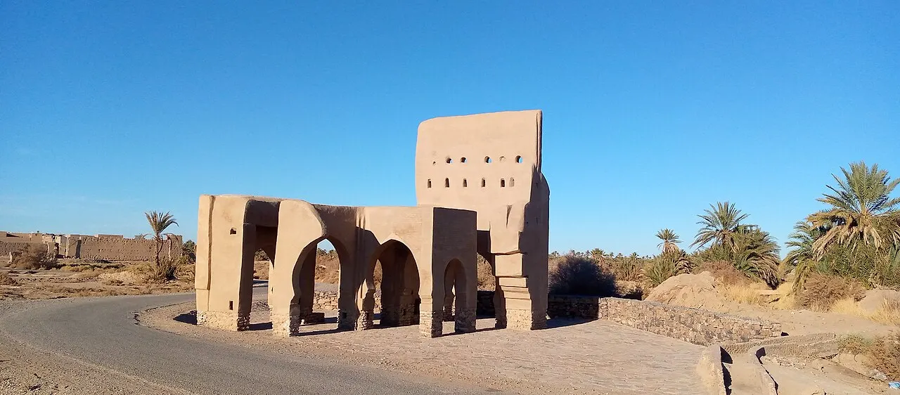 Portão norte de terra crua das ruínas de Sijilmassa, no Marrocos, com arcos e palmeiras ao fundo.