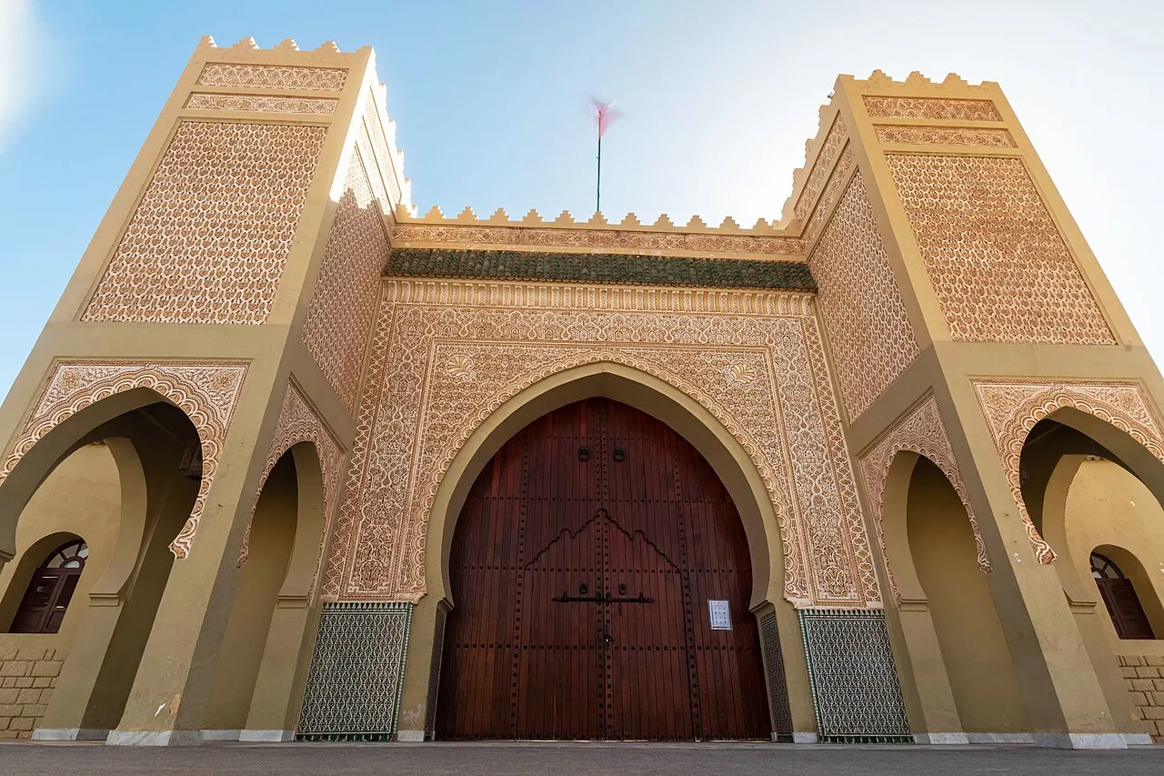 Portão monumental do mausoléu de Moulay Ali Cherif, em Rissani: arco em ferradura com porta dupla de madeira escura, estuque lavrado em toda a frente, azulejo verde no rodapé e duas torres ameadas.