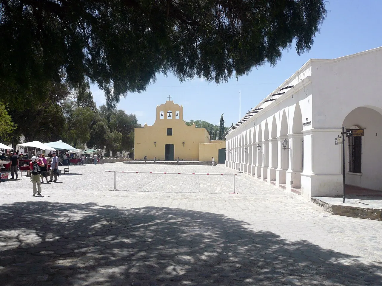 La plaza de Cachi, con suelo claro a la sombra de los árboles, la iglesia amarilla al fondo y casas blancas con arcadas a la derecha.