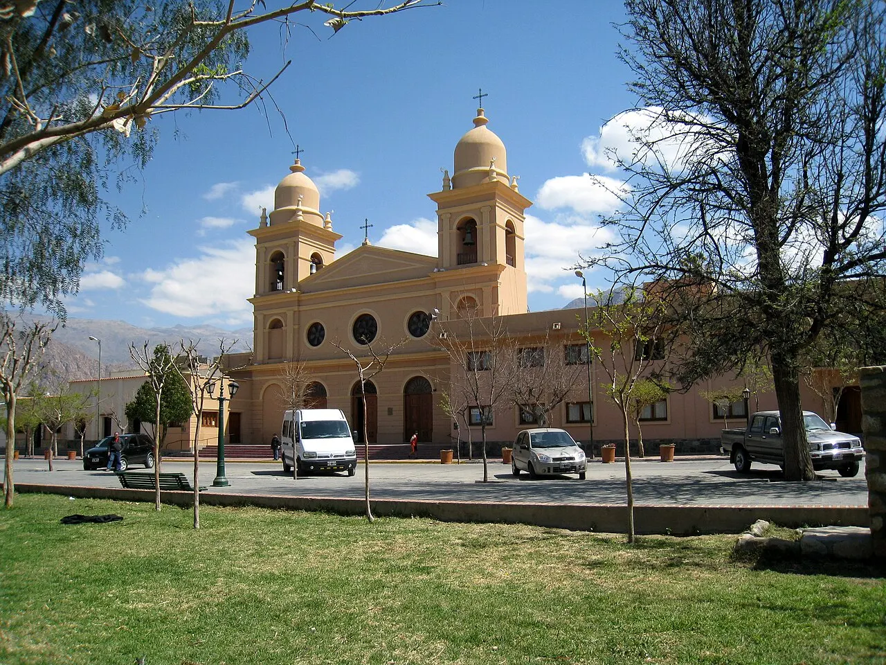 A catedral de Cafayate, de fachada amarela com duas torres e cinco naves, atrás da praça gramada e de uma rua com carros.