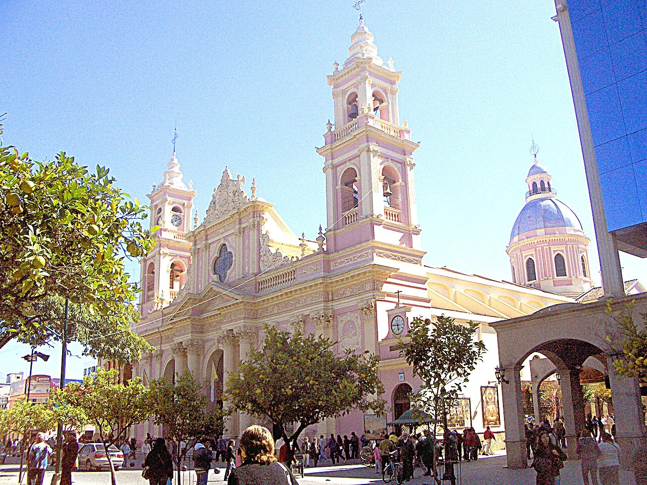 A Catedral Basílica de Salta, de fachada rosa e creme com duas torres e cúpula azul, vista da praça arborizada cheia de gente.