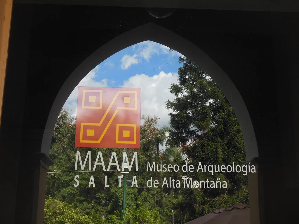 Inscrição do MAAM na porta envidraçada sob um arco pontudo, com árvores e céu refletidos no vidro, em Salta em 2013.