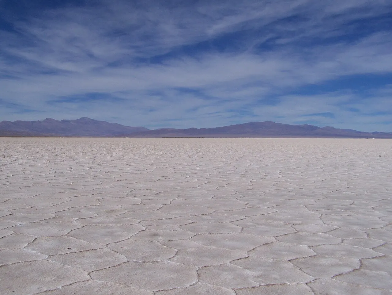 A planície branca das Salinas Grandes, com a crosta de sal rachada em hexágonos até o horizonte de montanhas, sob céu com nuvens finas.