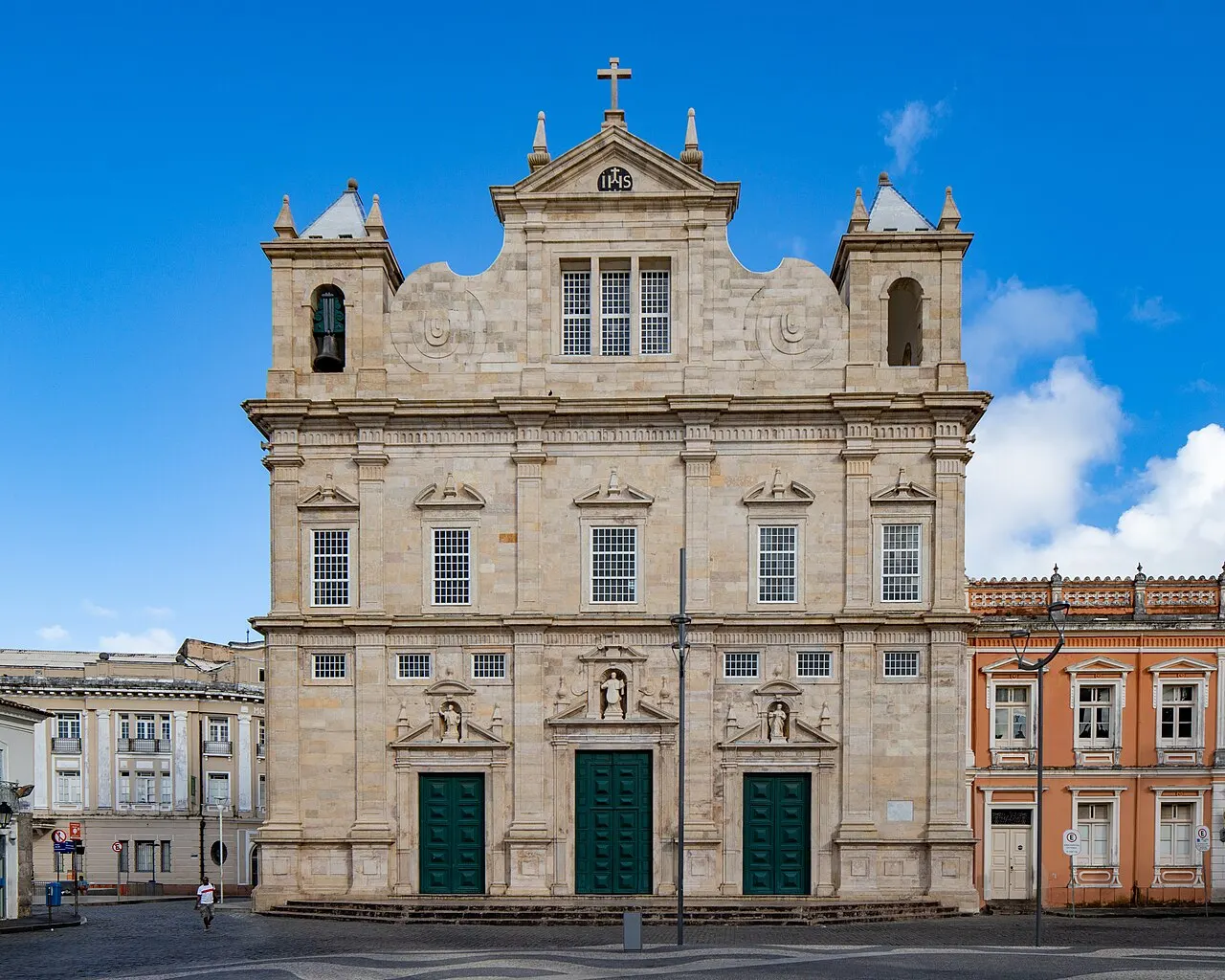 Fachada de pedra clara da Catedral Basílica de Salvador, com duas torres e portas verdes, sob céu azul.
