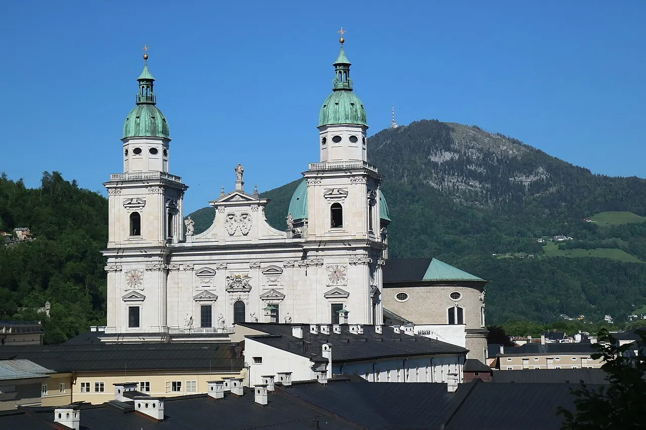 A fachada branca da Catedral de Salzburgo, com duas torres de cúpula verde, e o morro do Gaisberg atrás, sob céu azul.