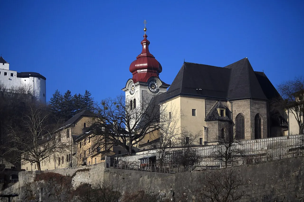 O Convento de Nonnberg, com a torre de cúpula vermelha em forma de bulbo e o telhado escuro da igreja, na encosta abaixo da fortaleza.