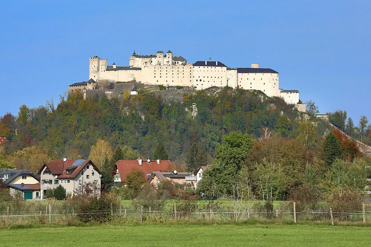A Fortaleza de Hohensalzburg, branca e comprida, no alto do morro arborizado, com casas e um prado na frente, sob céu azul.