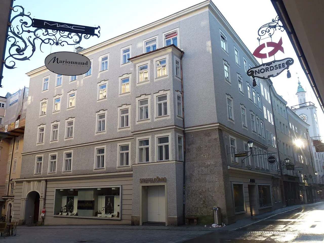 Uma fachada de cinco andares na Getreidegasse, com placas de ferro forjado penduradas sobre a rua estreita, sob céu azul.