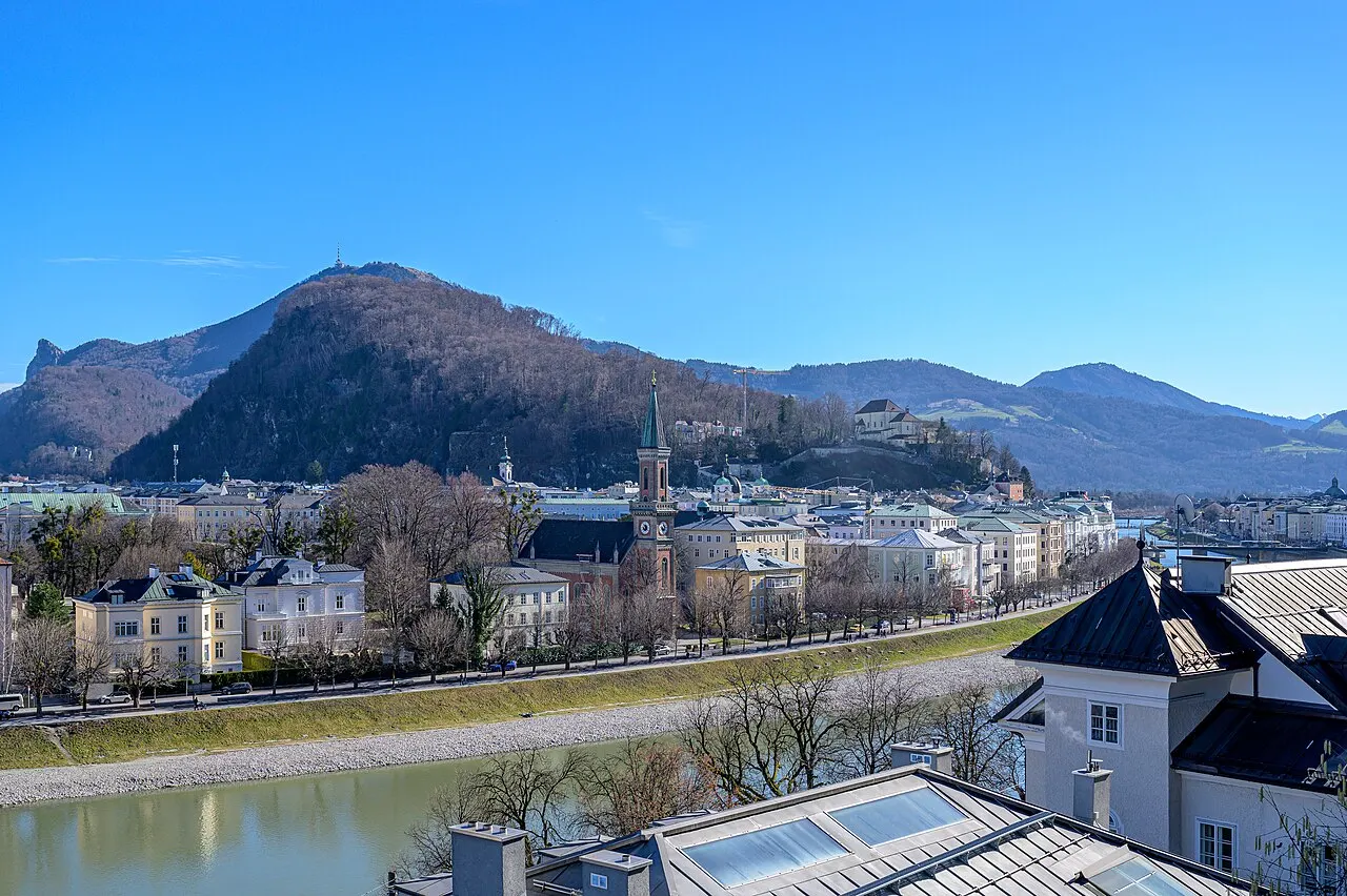O morro arborizado do Kapuzinerberg acima das casas da margem direita do Salzach, com montanhas atrás e céu azul.