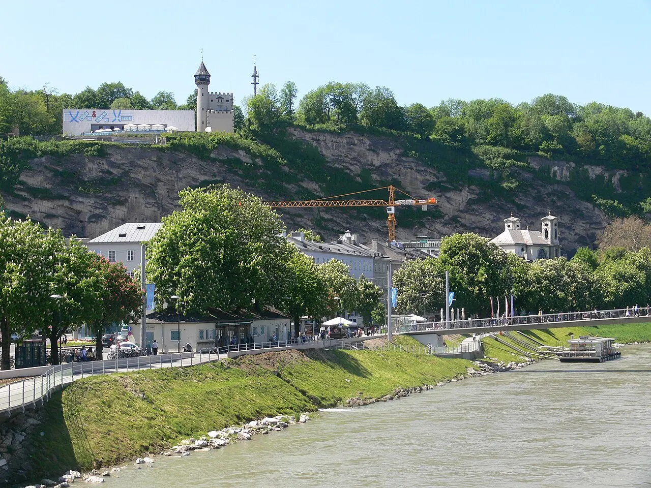 O paredão verde do Mönchsberg acima do rio Salzach e das margens gramadas, com o prédio branco do Museu de Arte Moderna no topo.