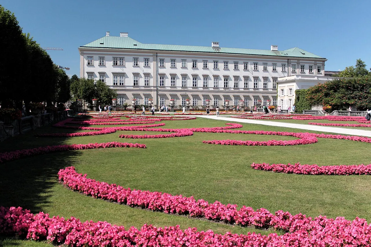 Os canteiros de flores rosas em arabesco do Jardim de Mirabell, com o palácio branco de Mirabell ao fundo e céu azul.