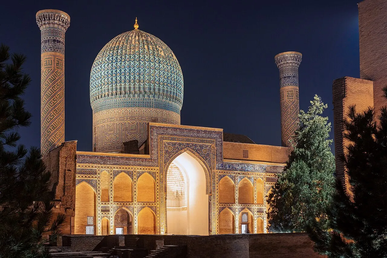 Mausoléu Gur-e-Amir, em Samarcanda, com a cúpula azul canelada e dois minaretes, iluminado à noite.