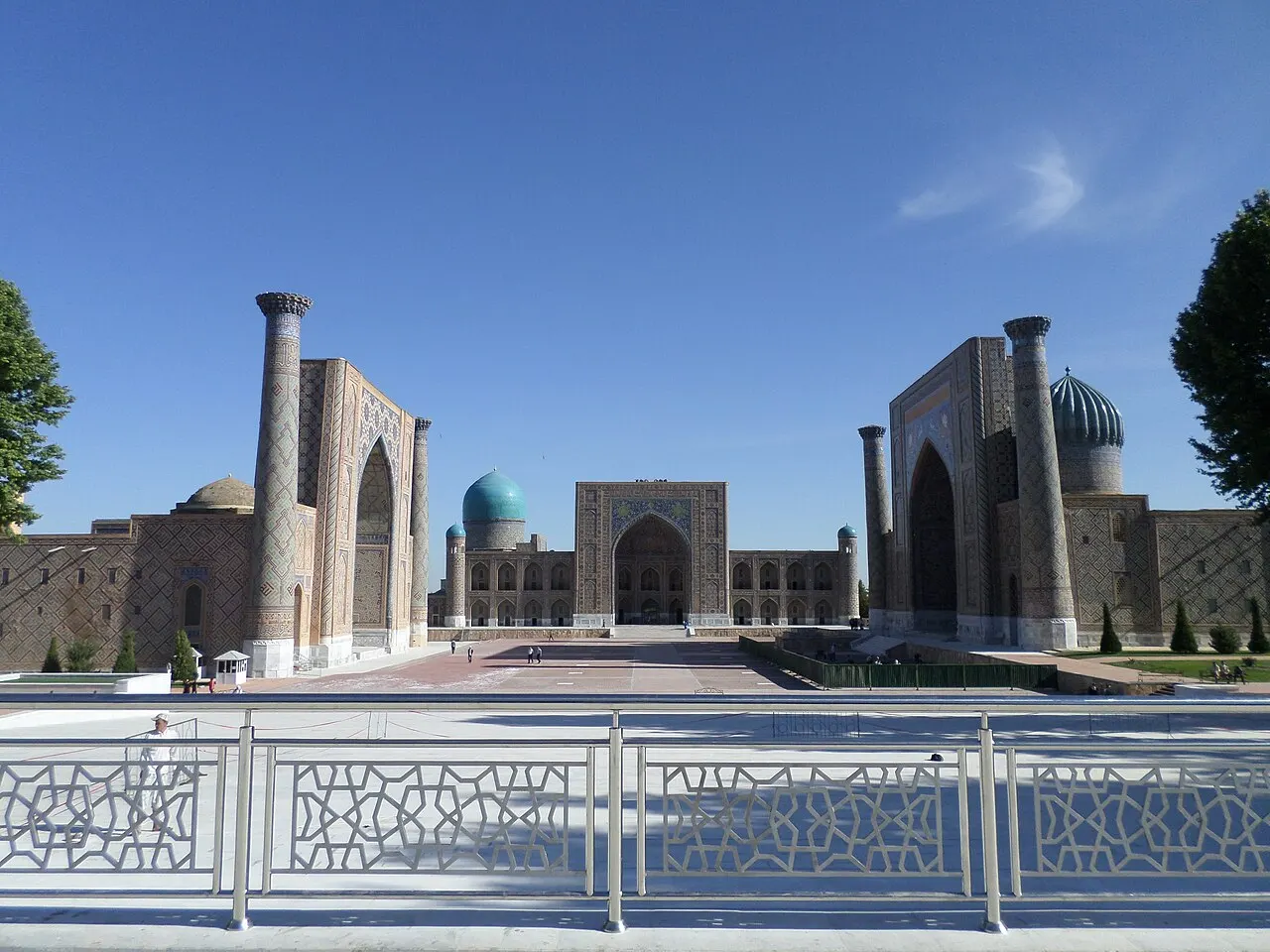 As três madraçais do Registan, em Samarcanda, com portais de azulejo e cúpulas azuis, vistas da borda da praça.