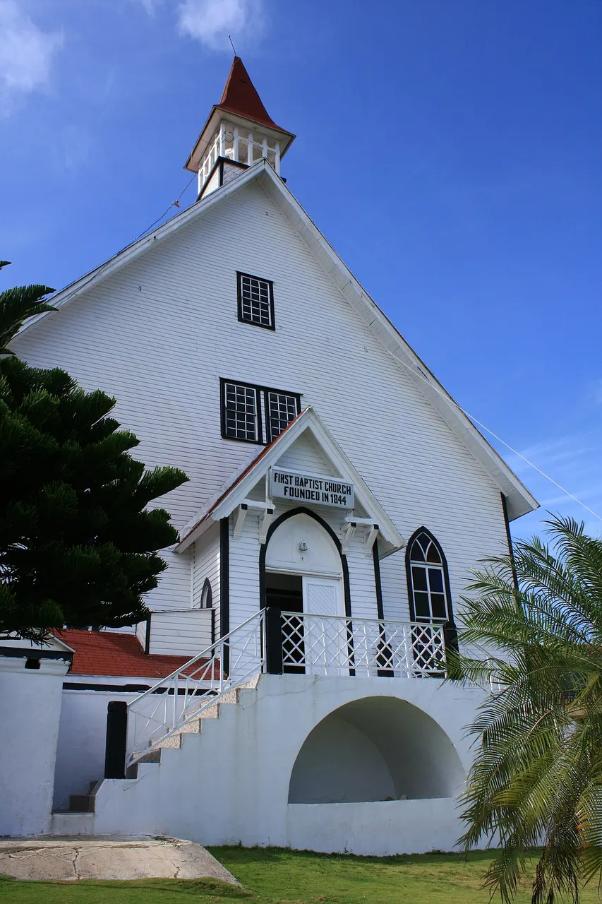 A First Baptist Church, em La Loma, uma igreja de madeira pintada de branco com torre pontuda vermelha, escadaria em arco e céu azul.