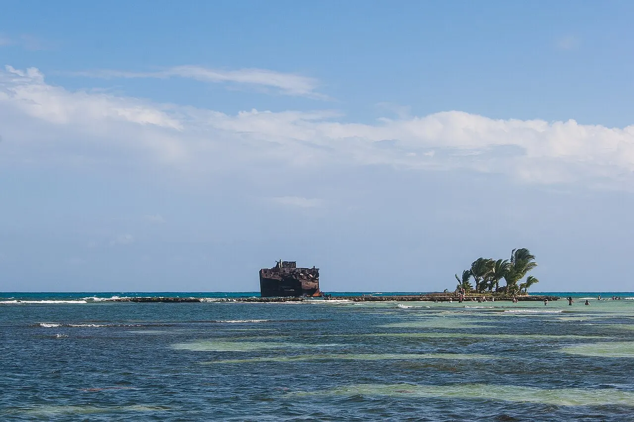 Rocky Cay ao longe, um ilhéu com coqueiros ao lado do casco enferrujado de um navio encalhado, sobre o recife raso.