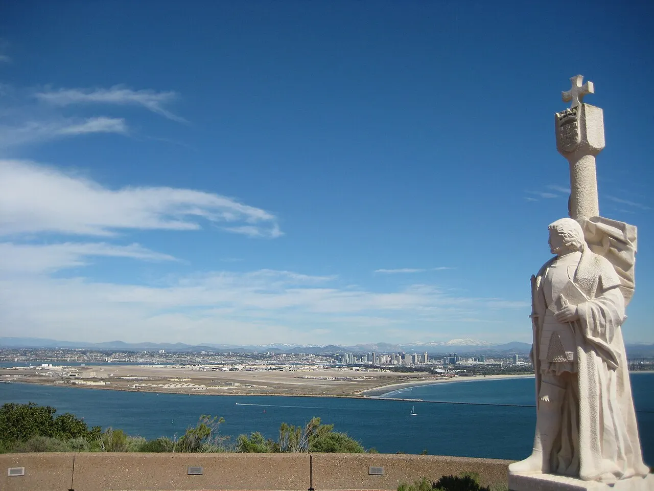 Estátua de pedra clara de um navegador diante da baía, com a cidade e a costa ao fundo sob céu azul