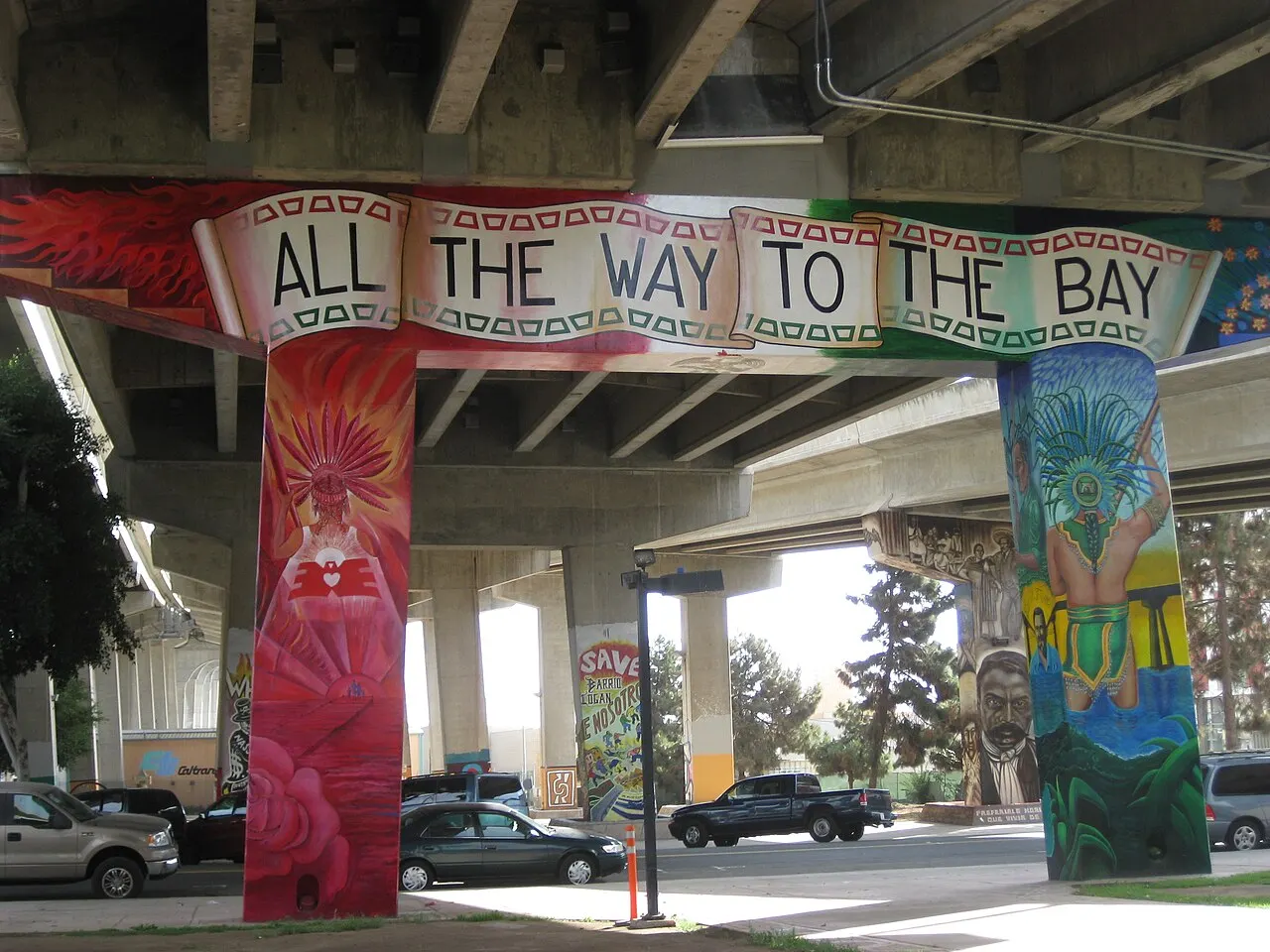 Pilares de viaduto pintados com murais coloridos e a faixa «All the Way to the Bay» sob a pista de concreto