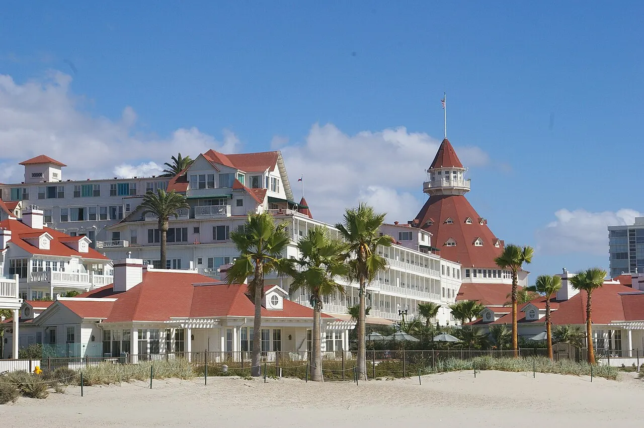Hotel vitoriano branco de telhados vermelhos e torre cônica, com palmeiras e areia em primeiro plano