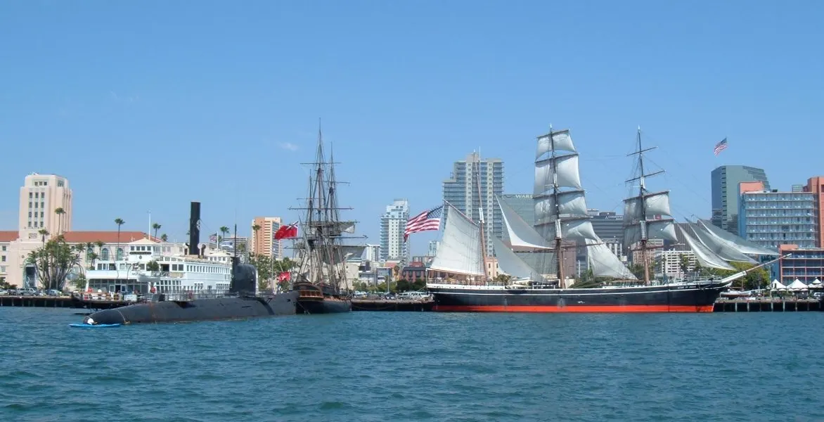 Veleiros antigos de mastros altos e um submarino atracados na baía, com prédios do centro ao fundo